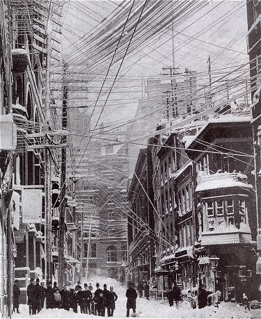 A group of people stand in the street in the aftermath of a large blizzard in New York City 1888. Power lines criss-cross the sky.