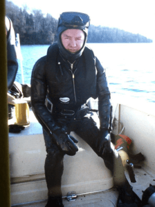 A man sits on the edge of a boat wearing a black scuba suit