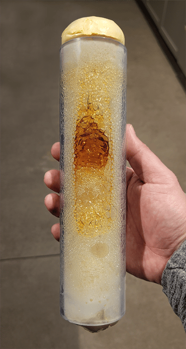 A hand holding a 8-inch piece of clear conduit. Inside the conduit is a 1-part spray foam that is cured on the ends, but uncured and looks like syrup in the middle of the seal.