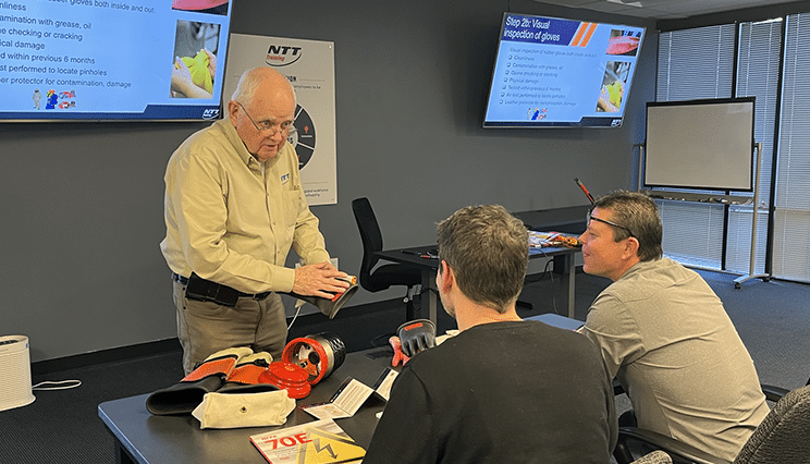 In a classroom with some tables and a couple TVs, a male teacher with glasses holds a pair of ppe rubber gloves while instructing two male electrical trainees on how to inspect them for damage.