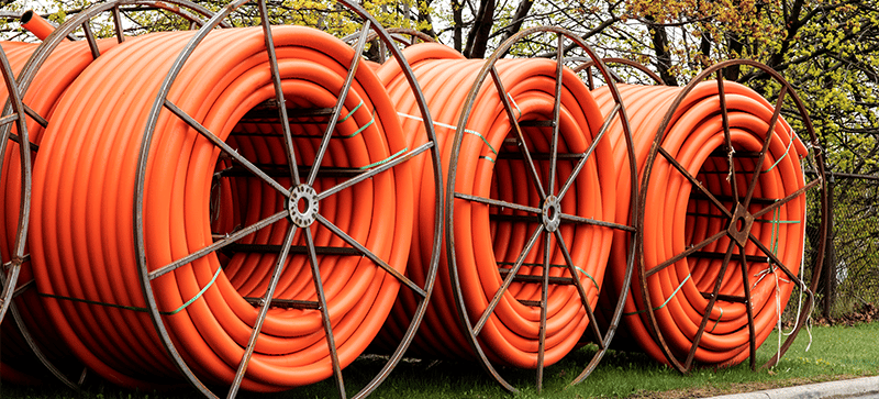 Trois grandes bobines de conduit de câble de télécommunications orange sont posées sur de l'herbe, avec des arbres en arrière-plan.