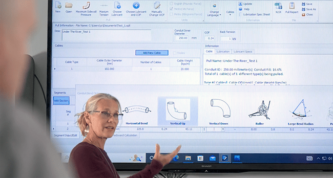 Eine blonde Frau mit Brille steht vor einem großen Fernsehbildschirm und hält eine Präsentation über die Software auf dem Bildschirm.