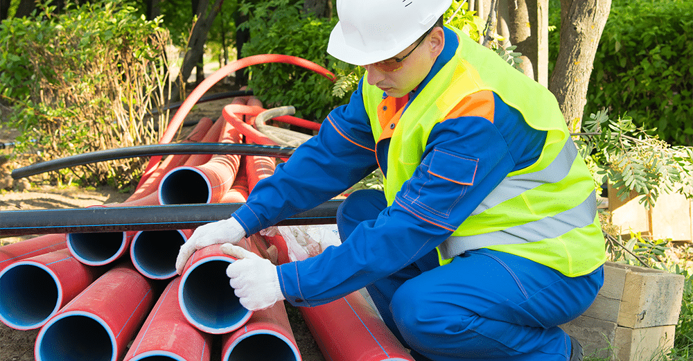 A worker in a white construction hat and green safety vest crouches down to inspect a pile of large red, empty conduits.
