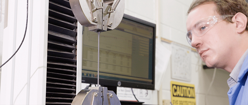 A scientist observes a test of the elongation of a conduit sample. The plastic strip is pulled from both ends by two gripping clamps.