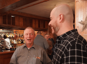 A smiling elderly man makes a younger man in the foreground laugh.