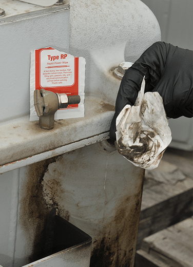 A hand holds a filthy wipe after having used it to clean grime off of some electrical equipment. The single-use pouch wrapper sits atop the equipment.