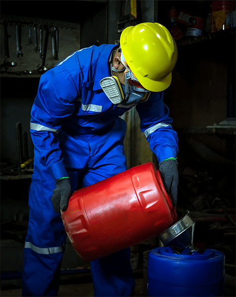 A man in a full body protective suit and a respirator pours a red pail of solvent into a funnel and a blue pail.
