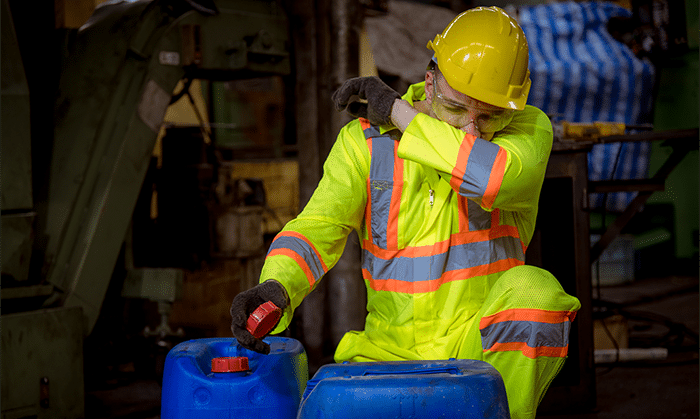 A worker in full safety gear, covers his noes and mouth as he opens a jug of solvent.
