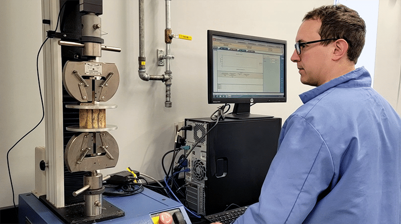 A scientist stands in front of a computer and monitors a machine that is crushing a foam block between two metal plates.