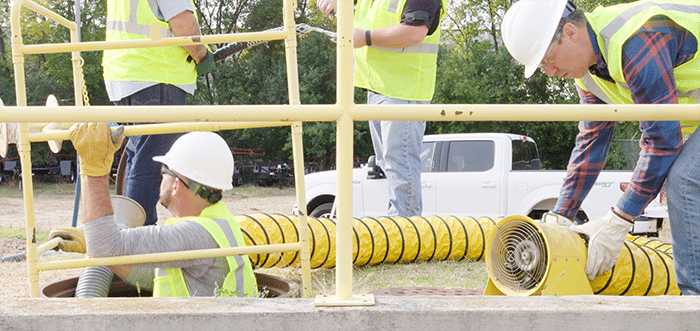 A man climbing into a manhole while another man prepares a yellow ventilation hose and fan. In the background is a white pickup truck and two more men preparing a cable to be pulled into the manhole.