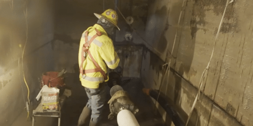 A worker in a yellow safety vest and a hard hat stands in an underground vault and examines a large high-voltage cable as it is pulled into a conduit opening.