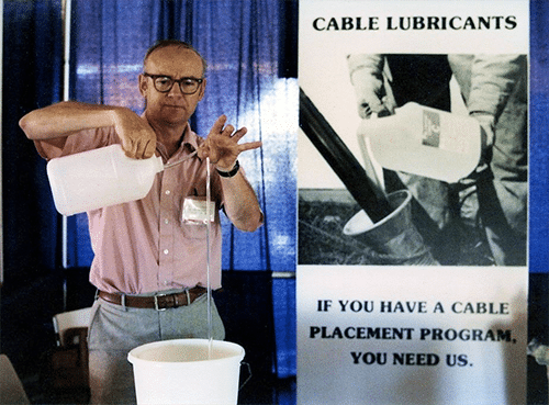 A man in glasses, a pink button up shirt, and slacks stands in his trade show booth with a sign that reads, "Cable Lubricants. If you have a cable placement program, you need us." The man is pouring a clear string of liquid from a jug into a pail from a height of 2 feet.