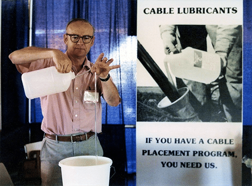 Un hombre con gafas, una camisa rosa con botones y pantalones de vestir se encuentra en su stand de feria con un cartel que dice: "Lubricantes para cables. Si tiene un programa de colocación de cables, nos necesita". El hombre vierte un hilo transparente de líquido de una jarra en una cubeta desde una altura de 2 pies.