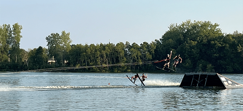 Members of the Midwest Ski Otter Waterskiing team jump of a ramp while holding a bottle of Polywater J-35 lubricant up in the air.