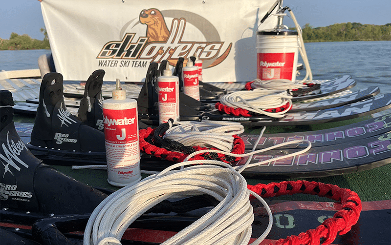 Water skis laid in a row on a dock. Each pair of skis has a red and white quart bottle of Polywater J lubricant. A white 5-gallon pail of Polywater J can be seen in the background next to a banner with a cartoon seal that reads "Ski Otters, Water Ski Team".