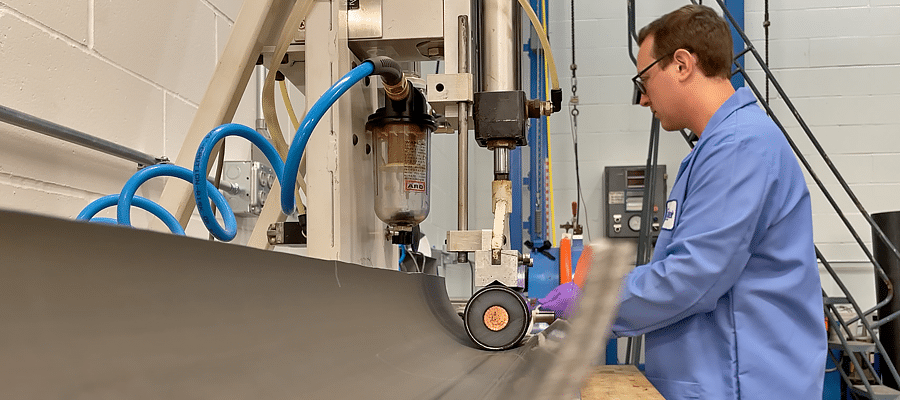 Un joven científico con gafas y una bata de laboratorio azul observa la mesa de pruebas de fricción de Polywater. Un trozo de conducto curvo tiene una sección corta de cable eléctrico de alto voltaje. Un mecanismo aplica presión al cable mientras se desliza a través del conducto.