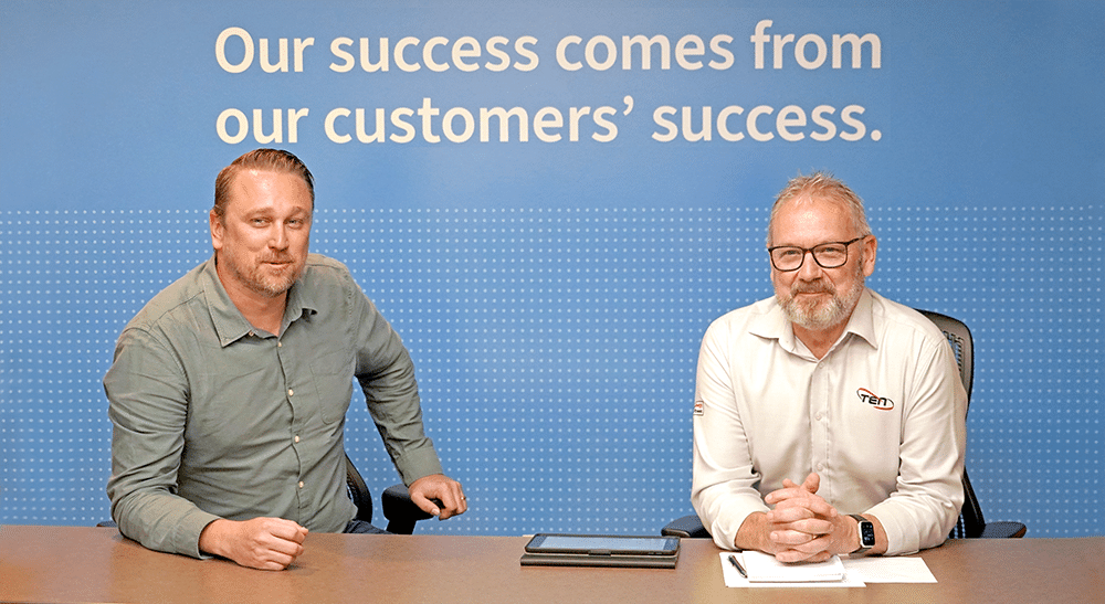 Two men sit at a table with a blue wall behind them with the text, "Our success comes from our customers' success." The man on the left, who has blond, slicked-back hair and a blond beard, is wearing a green, button-up shirt. The man on the right, who has greyish hair and a grey/white beard and glasses, is wearing a white, button-up shirt with the logo for "Ten Group" on his left chest.