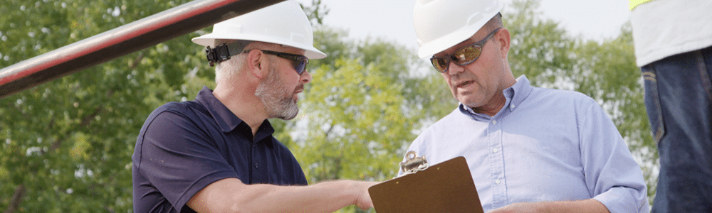 Zwei Männer mit weißen Schutzhelmen und Sonnenbrillen besprechen auf einer Baustelle zum Einziehen von Elektrokabeln einige Notizen auf einem Klemmbrett.