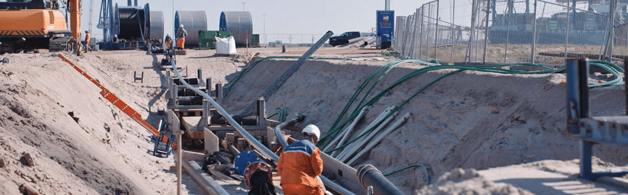 A worker in an orange jumpsuit and white hardhat, inspects a high-voltage electrical cable being pulled into a duct. The cable and worker are down in a large ditch about 200 feet long.