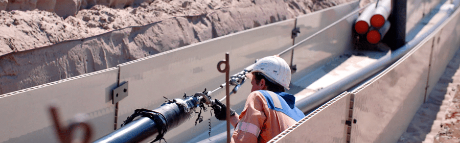 Ein Arbeiter in einem orangefarbenen Overall und einem weißen Schutzhelm untersucht eine Verbindung zwischen einem Stahleinzugskabel, das an ein Hochspannungskabel befestigt ist, das in einen Rohr gezogen wird.