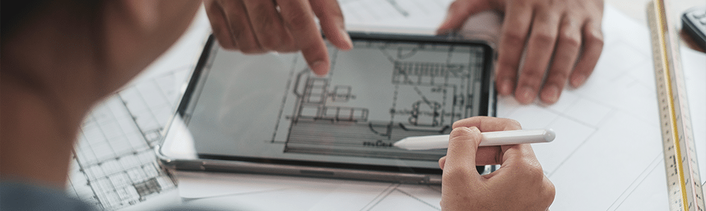 Una toma por encima del hombro de un ingeniero mientras mira un Ipad o tableta con algún tipo de diseño impreso en azul. El Ipad está sobre una mesa encima de unos planos de papel.