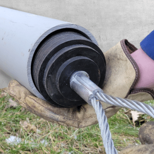 Une main gantée tient un conduit gris de 10 cm tout en insérant un épandeur de lubrifiant en caoutchouc noir d'où dépasse un câble d'acier en boucle.
