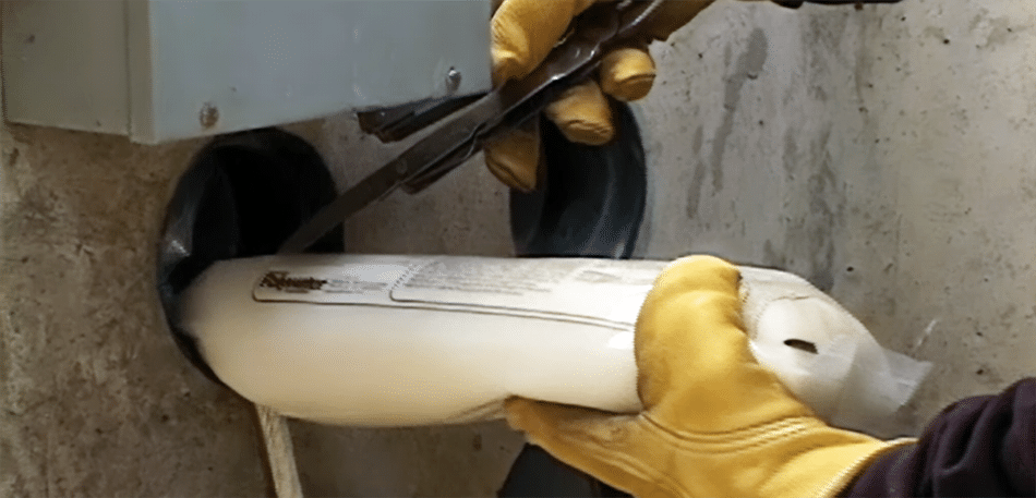 Dos manos enguantadas de color amarillo introducen un tubo de plástico lleno de lubricante Polywater J blanco en un banco de conductos en una pared de hormigón. La mano en la parte superior sostiene un cuchillo y corta la bolsa a medida que ingresa al orificio para prelubricar el ducto.