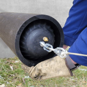 Une main gantée tient un grand conduit noir à l'intérieur duquel se trouve un bouchon noir gonflé en caoutchouc. Le bouchon est relié à une pince métallique, elle-même attachée à une corde qui sort de l'écran.