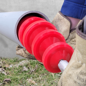 Une main gantée insère un épandeur de lubrifiant rouge à plusieurs disques dans un conduit gris qui sort d'un mur en béton.