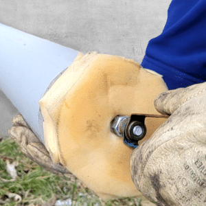 Hände mit Handschuhen halten ein großes graues Rohr, in das sie einen großen gelblichen Schwamm einführen. Im Schwamm befindet sich ein Bolzen und eine Metallschlaufe, an der ein Seil befestigt ist, um den Schwamm durch das Rohr zu ziehen.