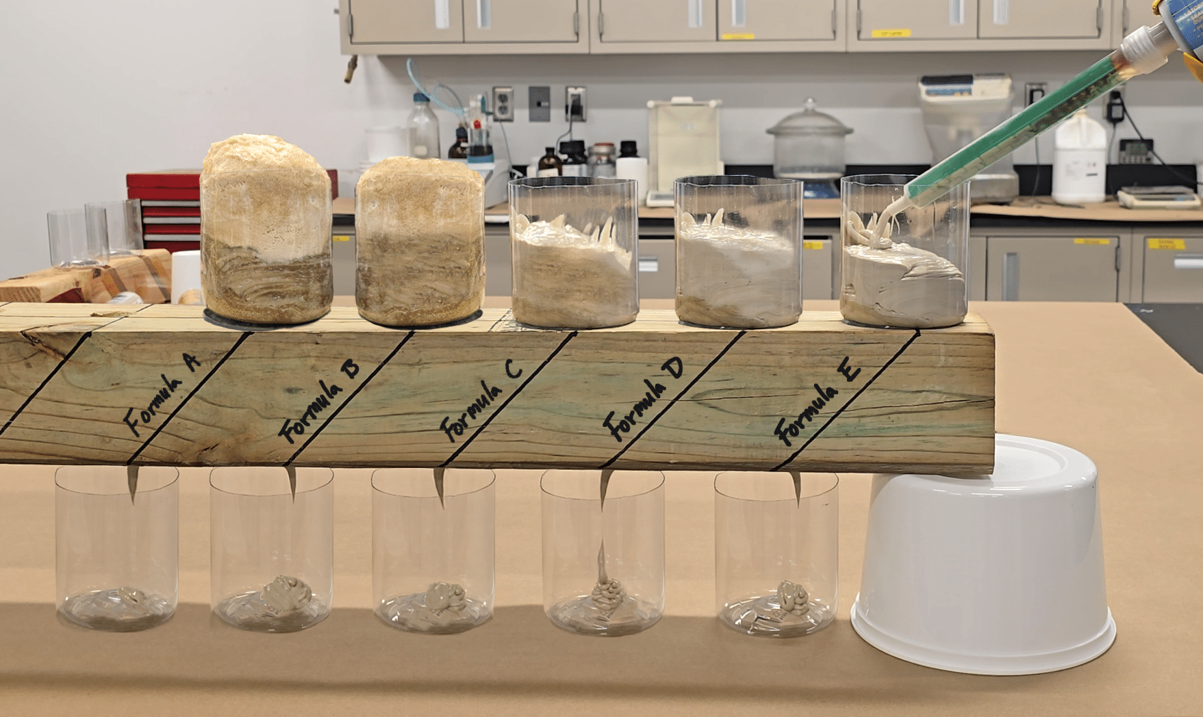 A 4x4 piece of wood with 5 cups on top of it filled with varying amounts of Polywater UPR. Below the board is 5 more cups to catch the excess sealant as it runs through the hole drilled in the board.