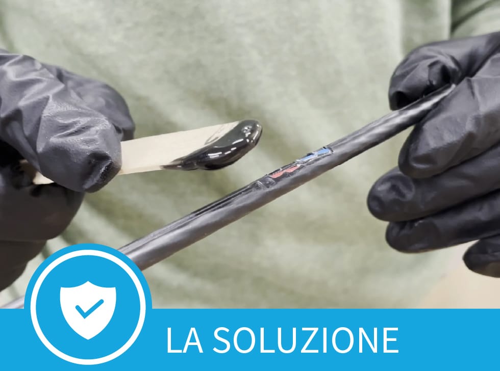 Una pasta nera - Polywater CJR Riparazione della Guaina del Cavo - sull'estremità di un bastoncino da gelato, viene spalmata sull'area danneggiata della guaina di un cavo in fibra. Nella parte inferiore dell'immagine si leggono le parole, 