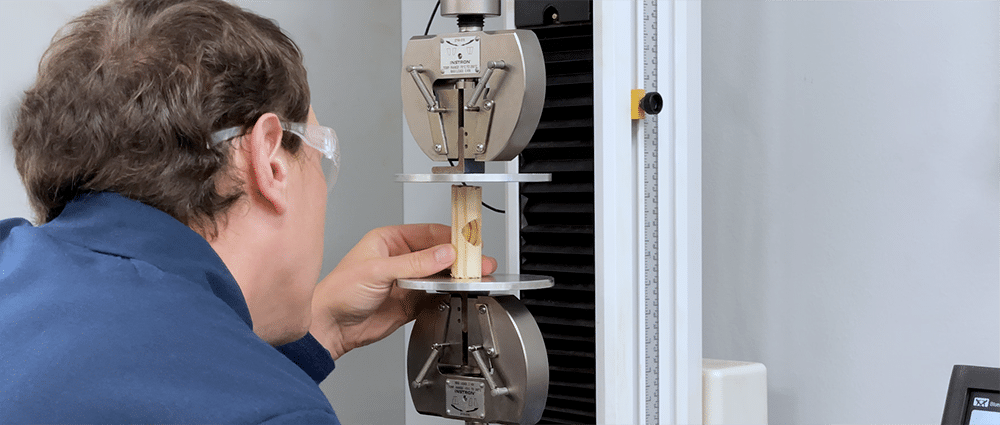 A scientist in a blue lab coat closely examines a small piece of a wooden dowel placed in a compression testing machine